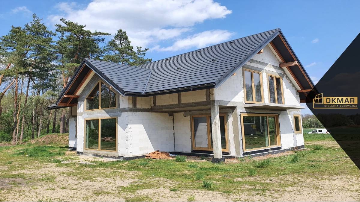 Budowa domu jednorodzinnego w stanie surowym otwartym z zamontowanymi oknami o drewnianej ramie, widoczne bloczki betonowe i dach z ciemnej dachówki ceramicznej.