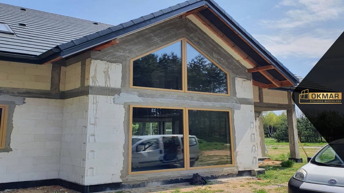 Surowy stan budynku z zamontowanymi oknami o drewnianej ramie, widoczne okno trójkątne na poddaszu i odbicie samochodu w dolnym oknie.