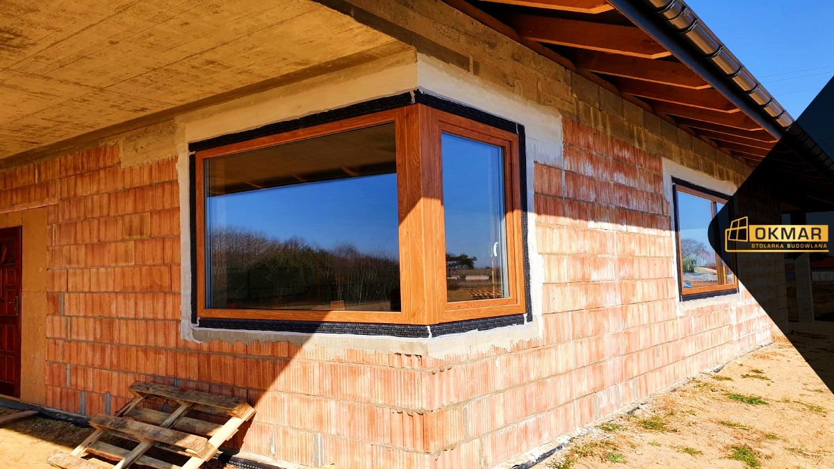 Montaż okna narożnego z drewnopodobną ramą w surowym budynku z czerwonej cegły, widoczna paleta i logo firmy w rogu zdjęcia.