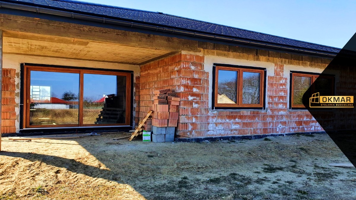Dom w trakcie budowy z zamontowanymi oknami i przesuwnymi drzwiami tarasowymi w drewnianej ramie, widoczne cegły i materiały budowlane.