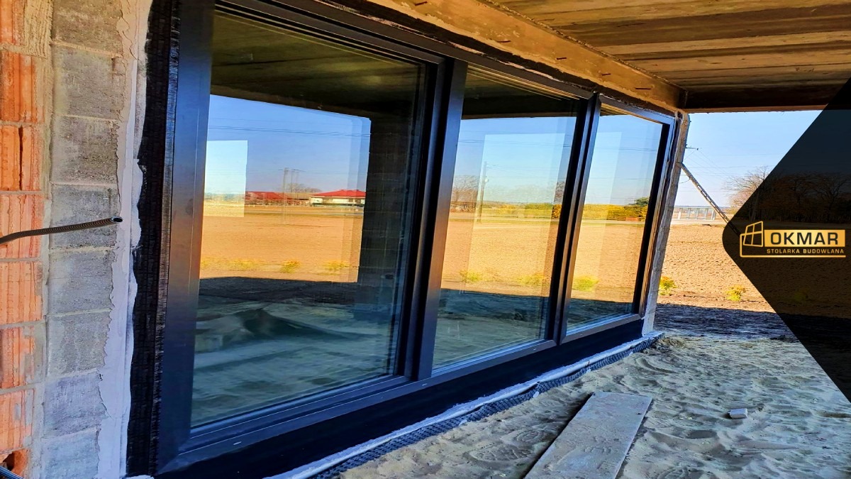 Montaż dużych, przesuwnych okien z czarnymi ramami w surowym stanie budynku, widoczna cegła i beton, na zewnątrz pole i dom.