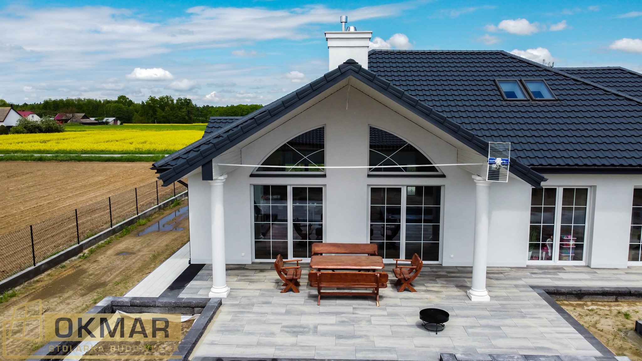 Nowoczesny dom z białymi kolumnami i dużymi oknami z białymi szprosami, widok z tarasu na pole rzepaku w oddali.