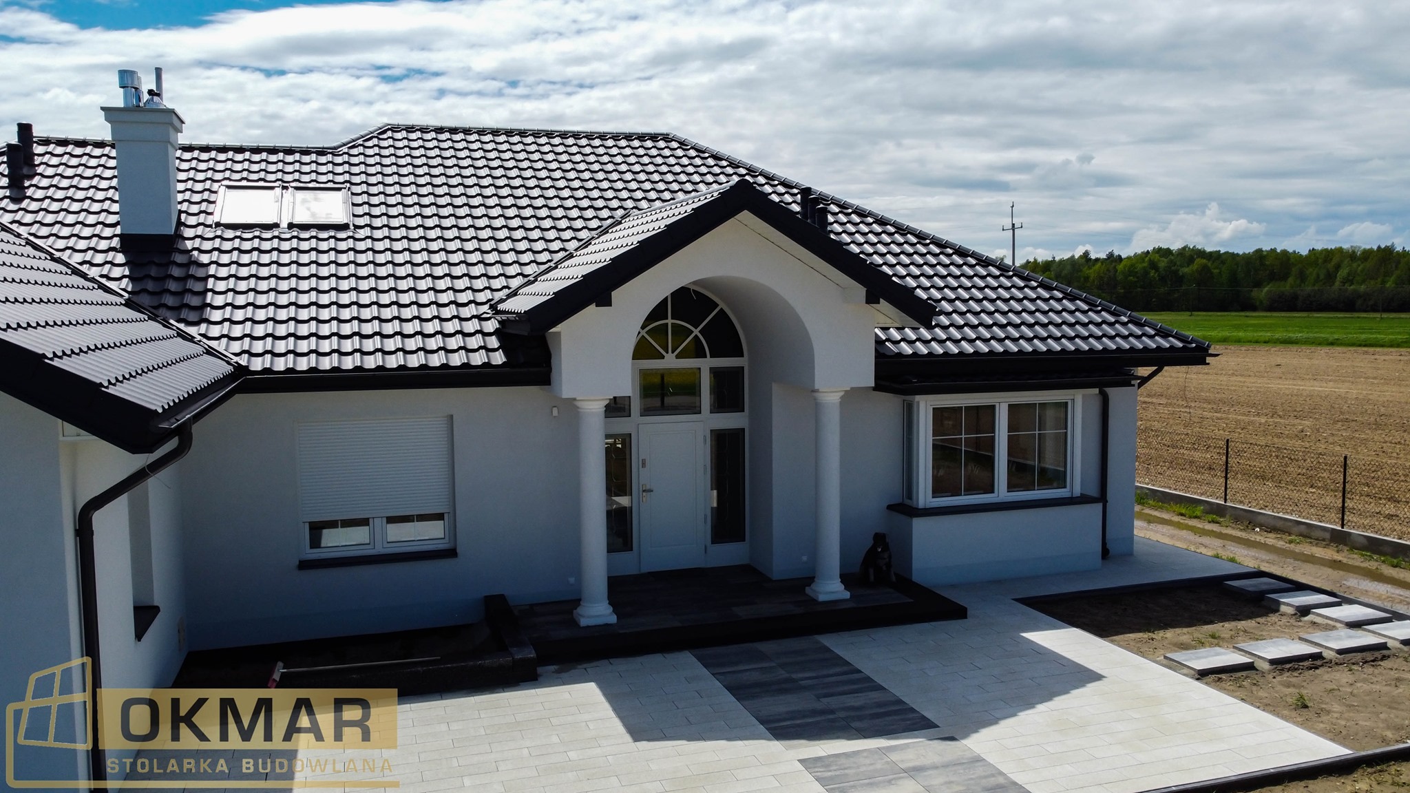 Nowoczesny dom jednorodzinny z czarnym dachem, białymi ścianami i stolarką okienną, widoczne okno z roletą zewnętrzną i wejście z kolumnami.