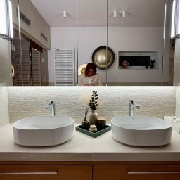 Autorska Pracownia Architektury i Wnętrz Gabriela Radwanowska - Nowoczesna łazienka z dwoma umywalkami nablatowymi, drewnianą szafką pod umywalką z subtelnym podświetleniem LED, lustrem na całej ścianie, w którym odbija się osoba robiąca zdjęcie telefonem.