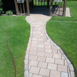 Kręta ścieżka z kostki brukowej prowadzi do drewnianej altany, otoczona soczystą zielenią trawnika. Wąż ogrodowy leży obok ścieżki.