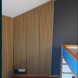 Ściana z paneli lamelowych w odcieniach beżu i czerni, zamontowana na skośnej ścianie w pokoju. Widoczna podłoga z paneli drewnianych i fragment ściany pomalowanej na ciemny kolor.