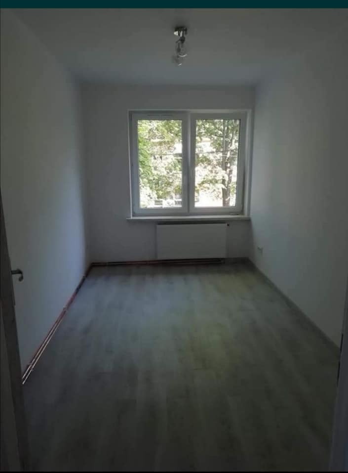 Jasne, puste pomieszczenie z szarą podłogą i białymi ścianami. Widoczne okno z widokiem na drzewa. Grzejnik pod oknem. Ujęcie z wnętrza pokoju.