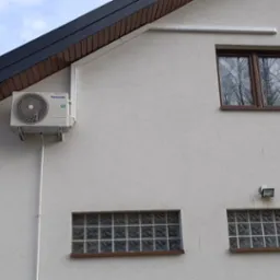 Biała jednostka zewnętrzna klimatyzatora Panasonic zamontowana na ścianie domu, widoczne okna i numer domu 32E.