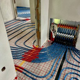 Paweł Łukasik SaniSafe Technika Grzewcza i Sanitarna - Instalacja ogrzewania podłogowego w Grybowie: niebieskie rurki na folii, rozdzielacz z zaworami w ścianie, widok z perspektywy podłogi, surowy stan wykończenia.