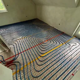 Instalacja ogrzewania podłogowego w poddaszu z oknami dachowymi. Rury w kolorze niebieskim ułożone na folii z czerwoną siatką i taśmą w kolorze żółtym.