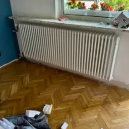 Biały, żeliwny grzejnik zamontowany pod oknem z kwiatami, na tle drewnianego parkietu i fragmentu niebieskiej ściany. Widoczne ślady po remoncie.