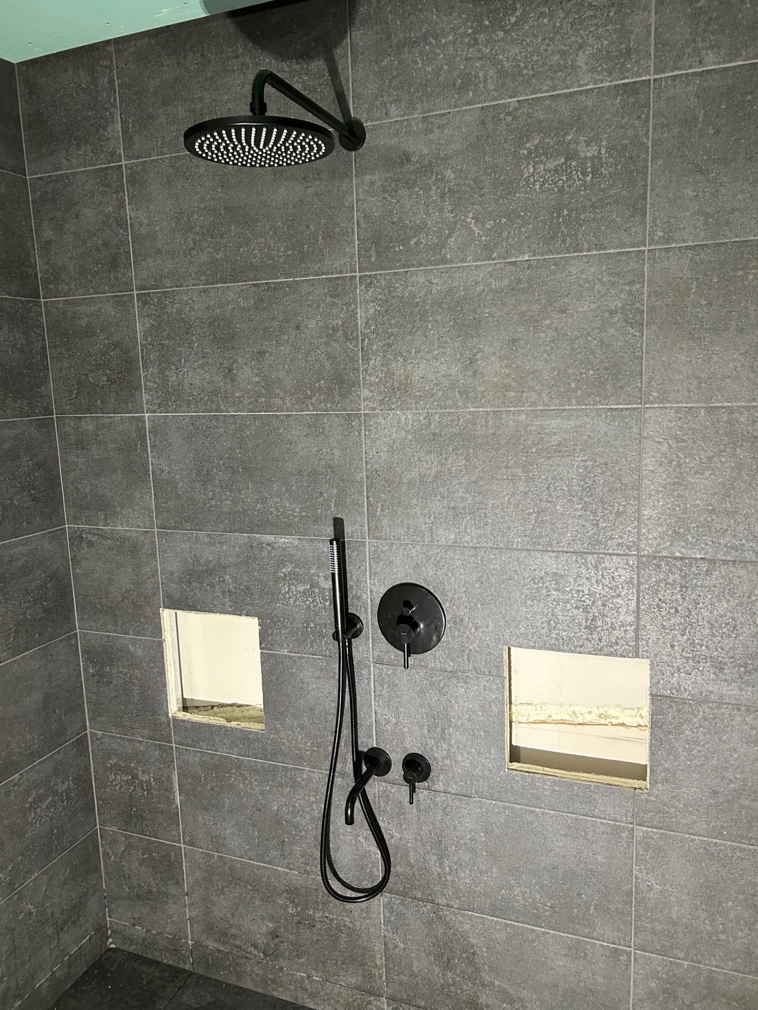 Szara, kwadratowa mozaika ścienna z czarną armaturą prysznicową i dwoma otworami w ścianie, ukazująca etap prac w łazience w Grybowie.