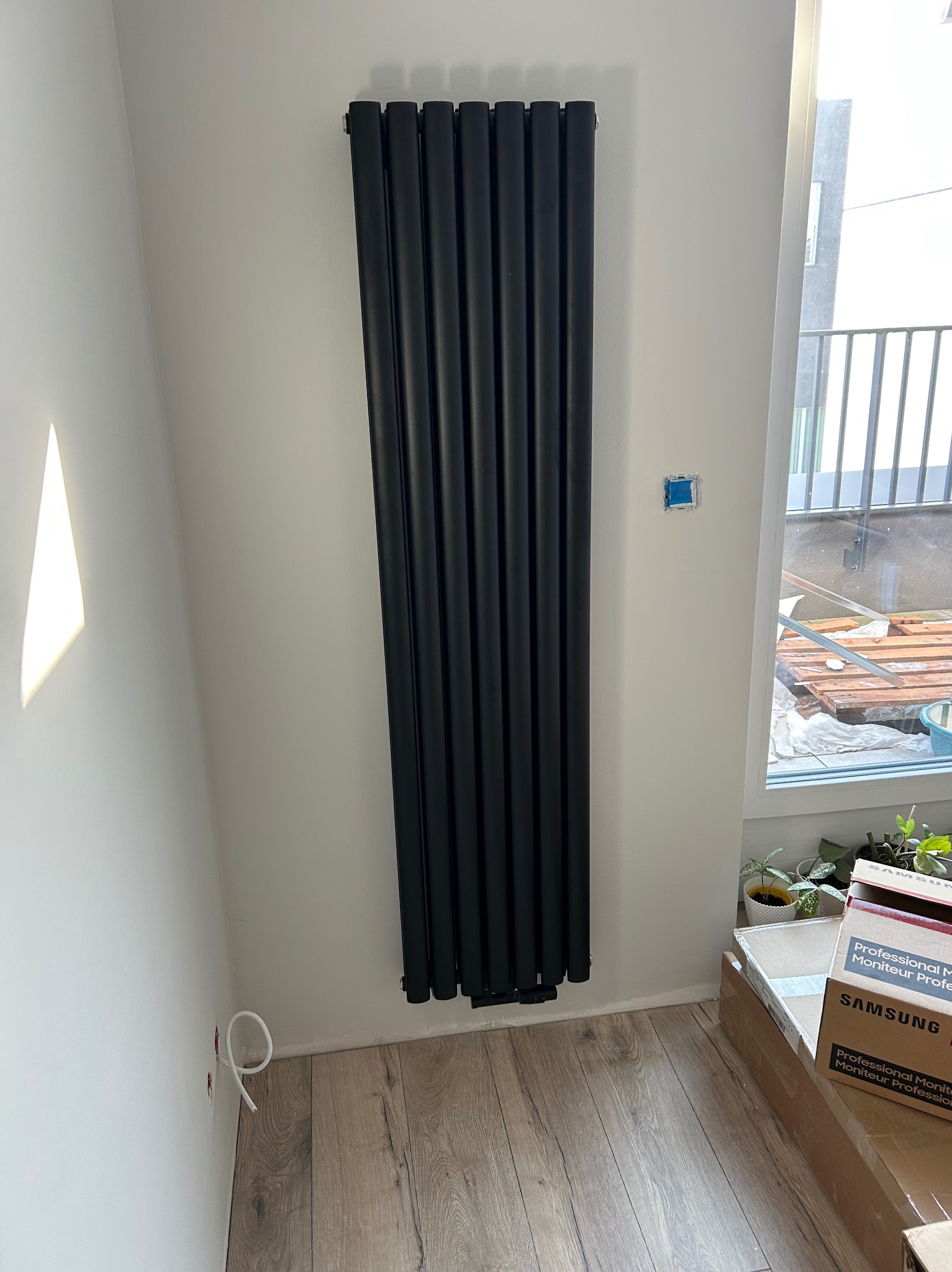 Wysoki, pionowy, czarny grzejnik panelowy zamontowany na białej ścianie w nowym budownictwie, widoczne wystające kable, podłączenie do instalacji w trakcie prac wykończeniowych.