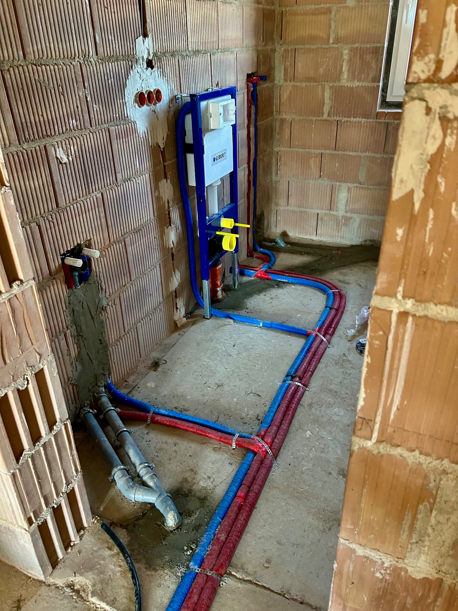 Instalacja wodno-kanalizacyjna w surowym wnętrzu: stelaż WC Geberit, rury w izolacji czerwonej i niebieskiej, przyłącza w ścianach z cegły. Prace hydrauliczne w toku.