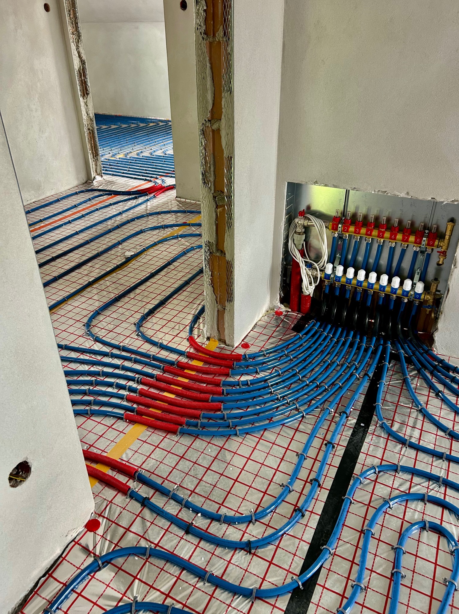 Instalacja ogrzewania podłogowego w Grybowie: niebieskie rurki na folii, rozdzielacz z zaworami w ścianie, widok z perspektywy podłogi, surowy stan wykończenia.