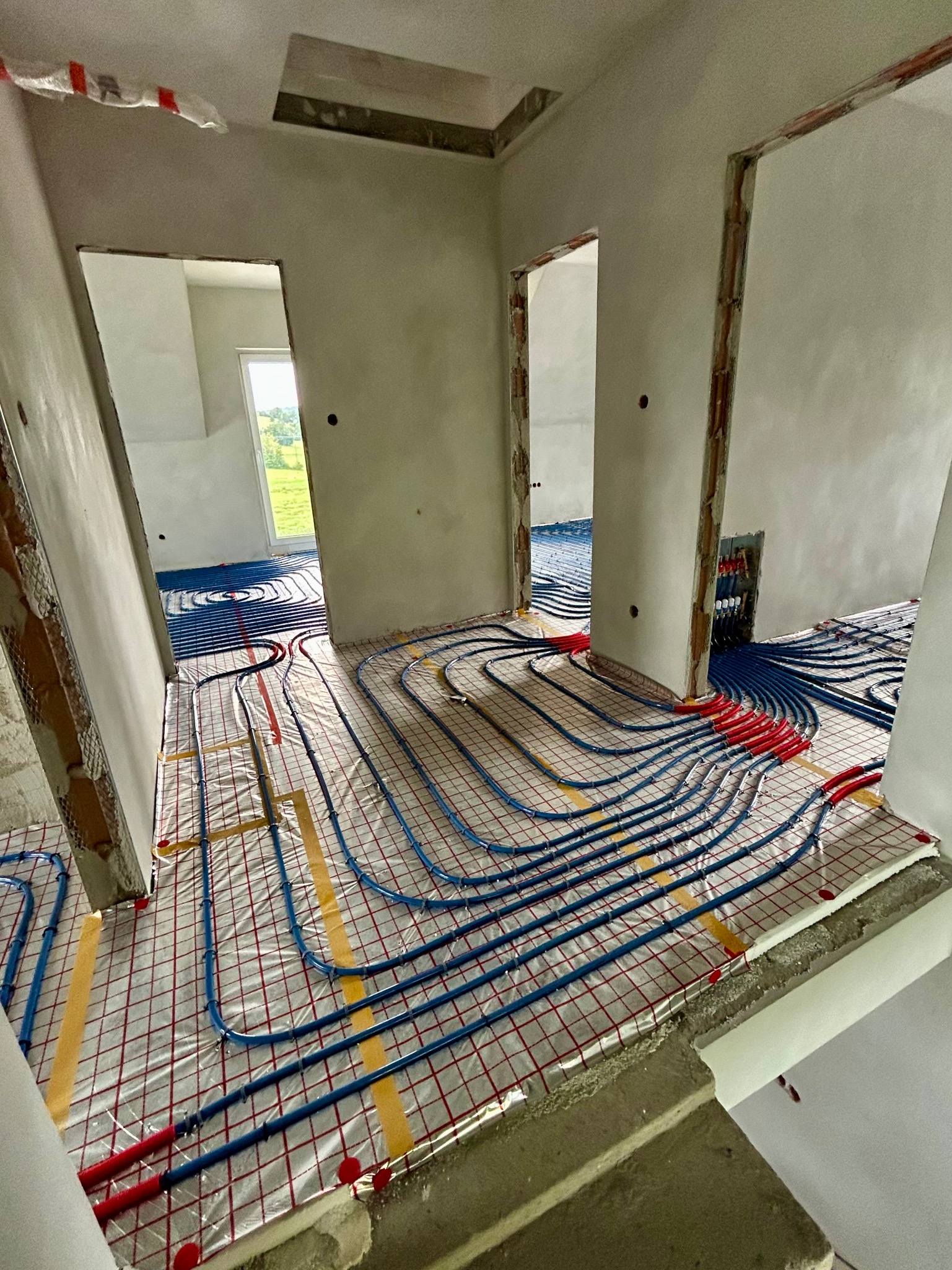 Niebieskie rury ogrzewania podłogowego ułożone na folii z siatką w surowym wnętrzu. Widoczne przygotowane przejścia do kolejnych pomieszczeń.