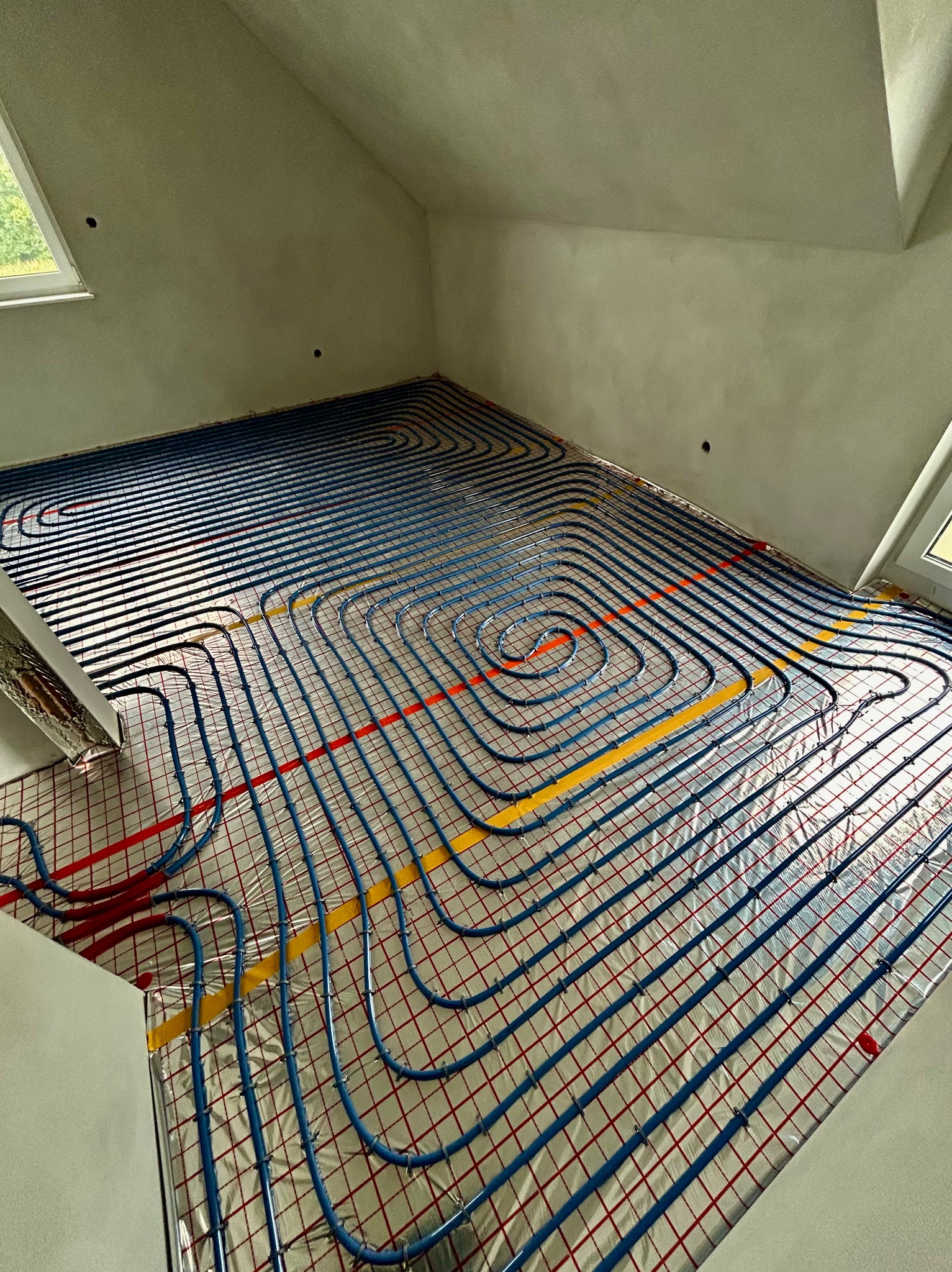 Instalacja ogrzewania podłogowego w poddaszu z oknami dachowymi. Rury w kolorze niebieskim ułożone na folii z czerwoną siatką i taśmą w kolorze żółtym.