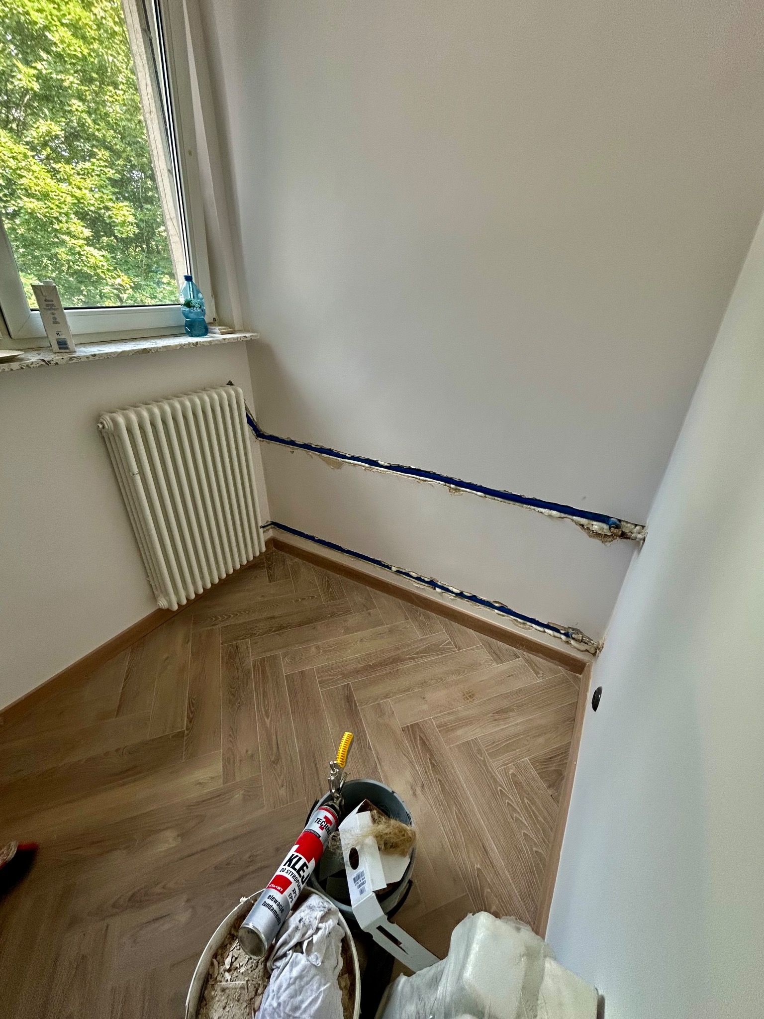 Wnętrze pokoju w trakcie prac hydraulicznych: widoczne rury w ścianie, grzejnik, podłoga w jodełkę i narzędzia pracy.