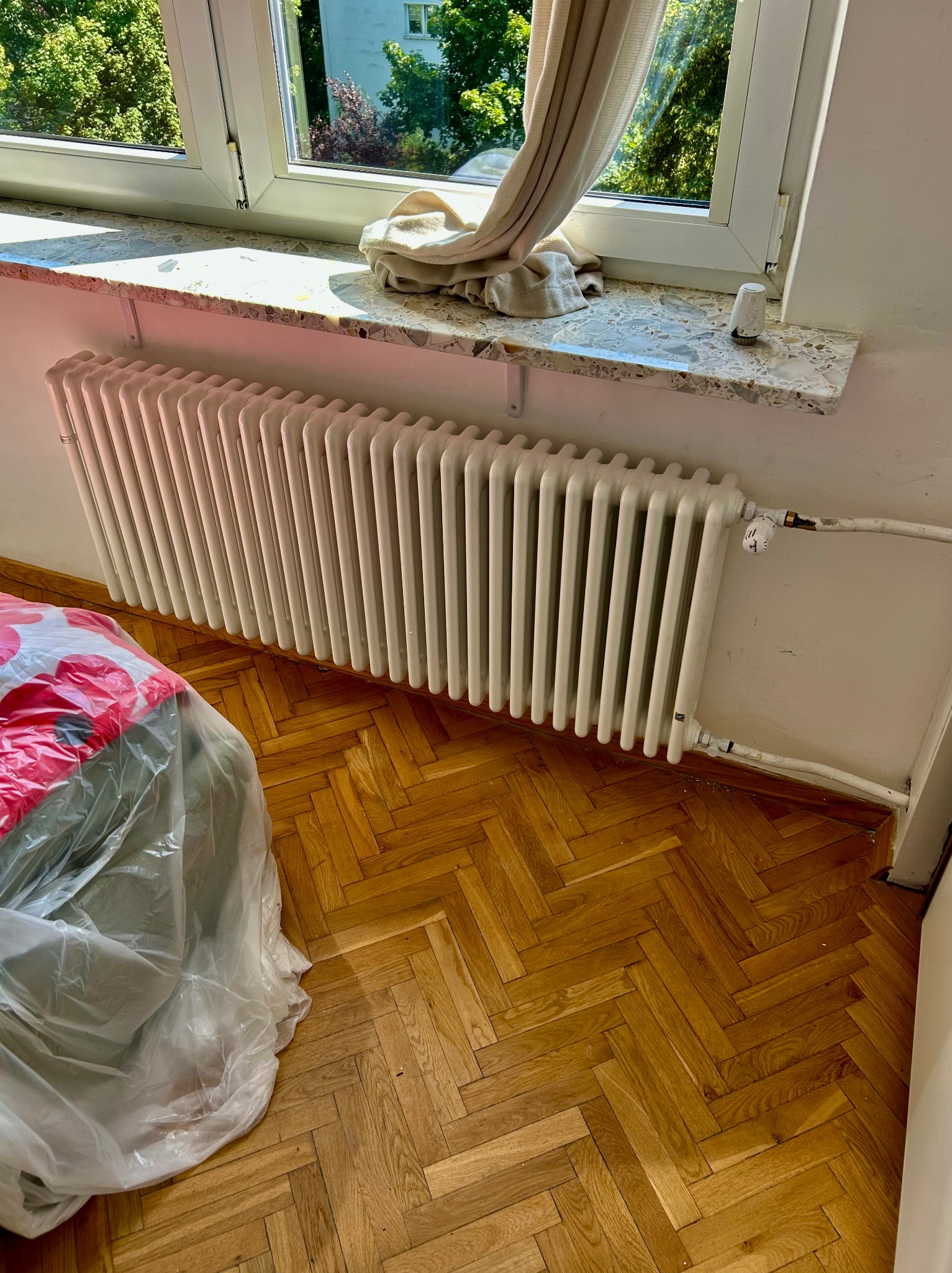 Biały, żeberkowy grzejnik zamontowany pod oknem z marmurowym parapetem. Na podłodze parkiet ułożony w jodełkę. Częściowo widoczne meble przykryte folią.
