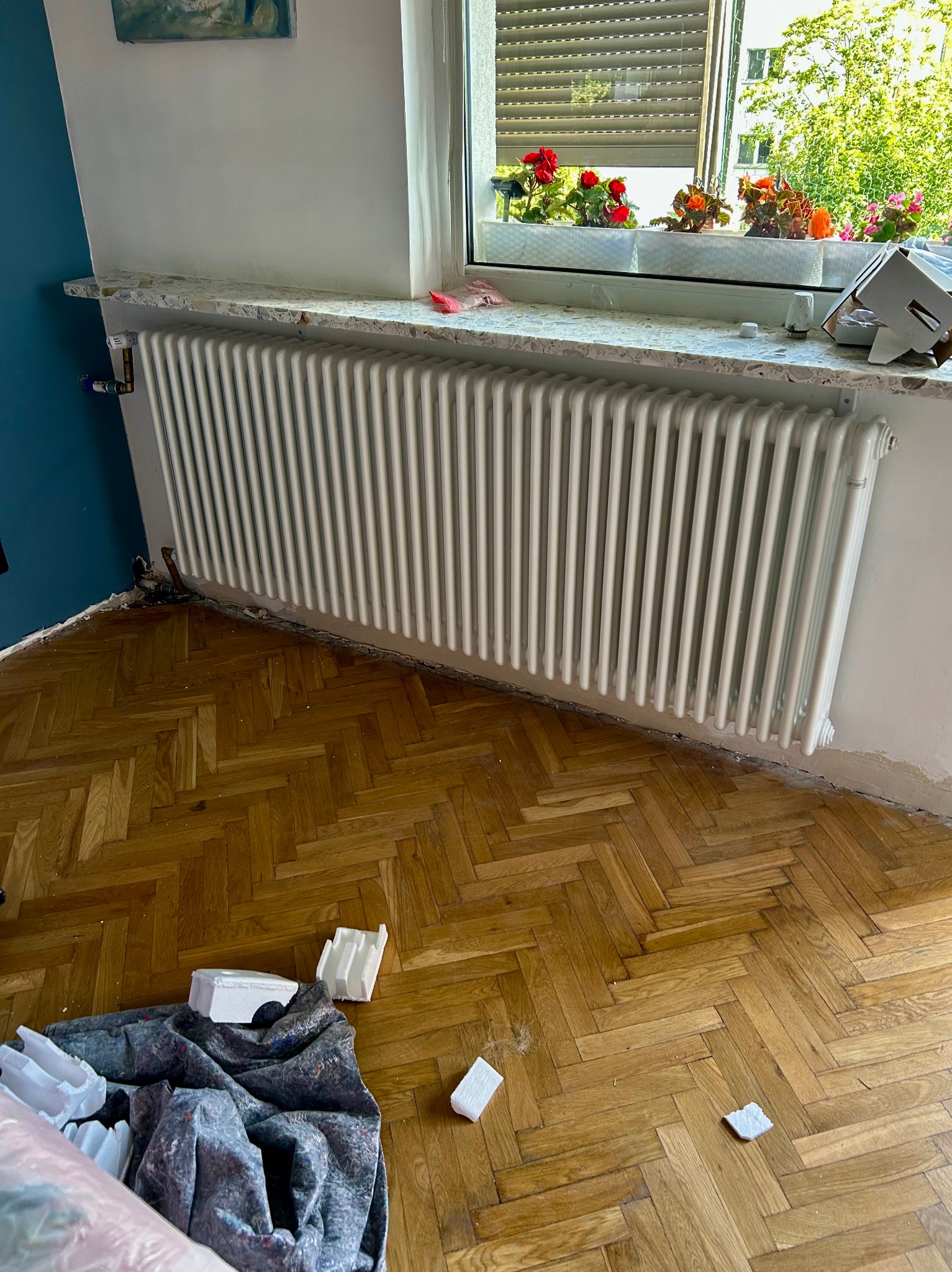 Biały, żeliwny grzejnik zamontowany pod oknem z kwiatami, na tle drewnianego parkietu i fragmentu niebieskiej ściany. Widoczne ślady po remoncie.