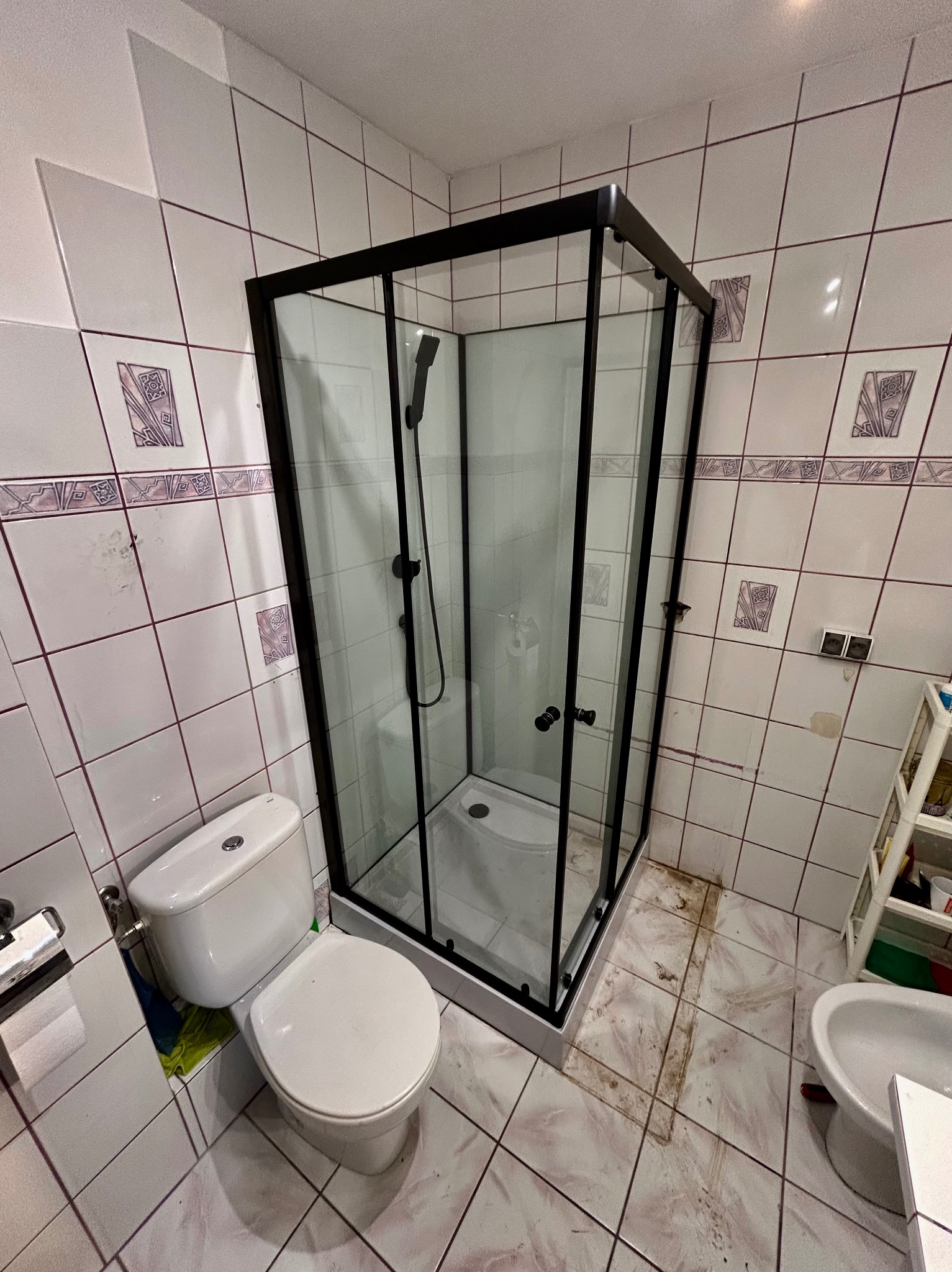 Nowoczesna kabina prysznicowa z czarnymi profilami w łazience z białą armaturą i wzorzystymi płytkami. Widok na toaletę i bidet.