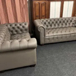 Komplet wypoczynkowy w stylu Chesterfield: sofa trzyosobowa i fotel, obite szarą skórą, z pikowaniem typu chesterfield, na tle brązowej szafy i wzorzystej zasłony.