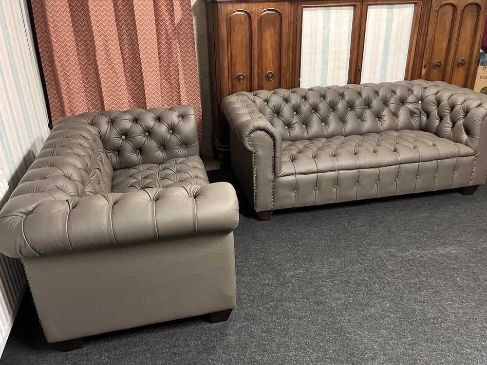 Komplet wypoczynkowy w stylu Chesterfield: sofa trzyosobowa i fotel, obite szarą skórą, z pikowaniem typu chesterfield, na tle brązowej szafy i wzorzystej zasłony.