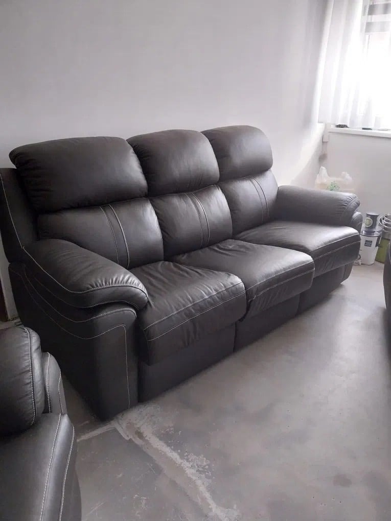 Ciemnobrązowa, skórzana sofa trzyosobowa z białymi przeszyciami, widoczna w salonie w Markach, z częściowo widocznym fotelem z tej samej serii.