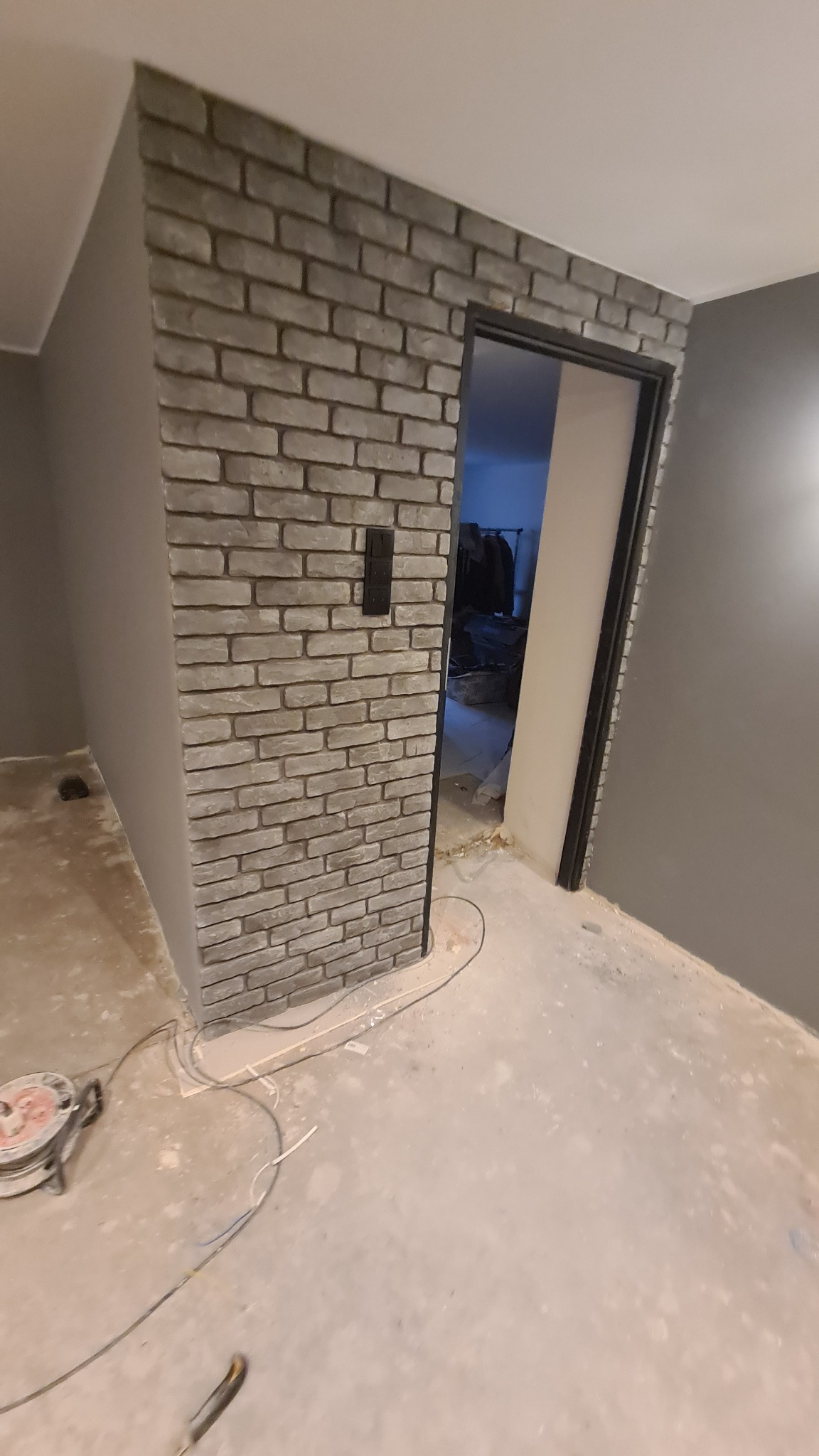 Fragment ściany wykończonej dekoracyjną cegłą w odcieniach szarości, z widocznym gniazdkiem elektrycznym i otwartym przejściem do kolejnego pomieszczenia, podłoga w trakcie prac wykończeniowych...