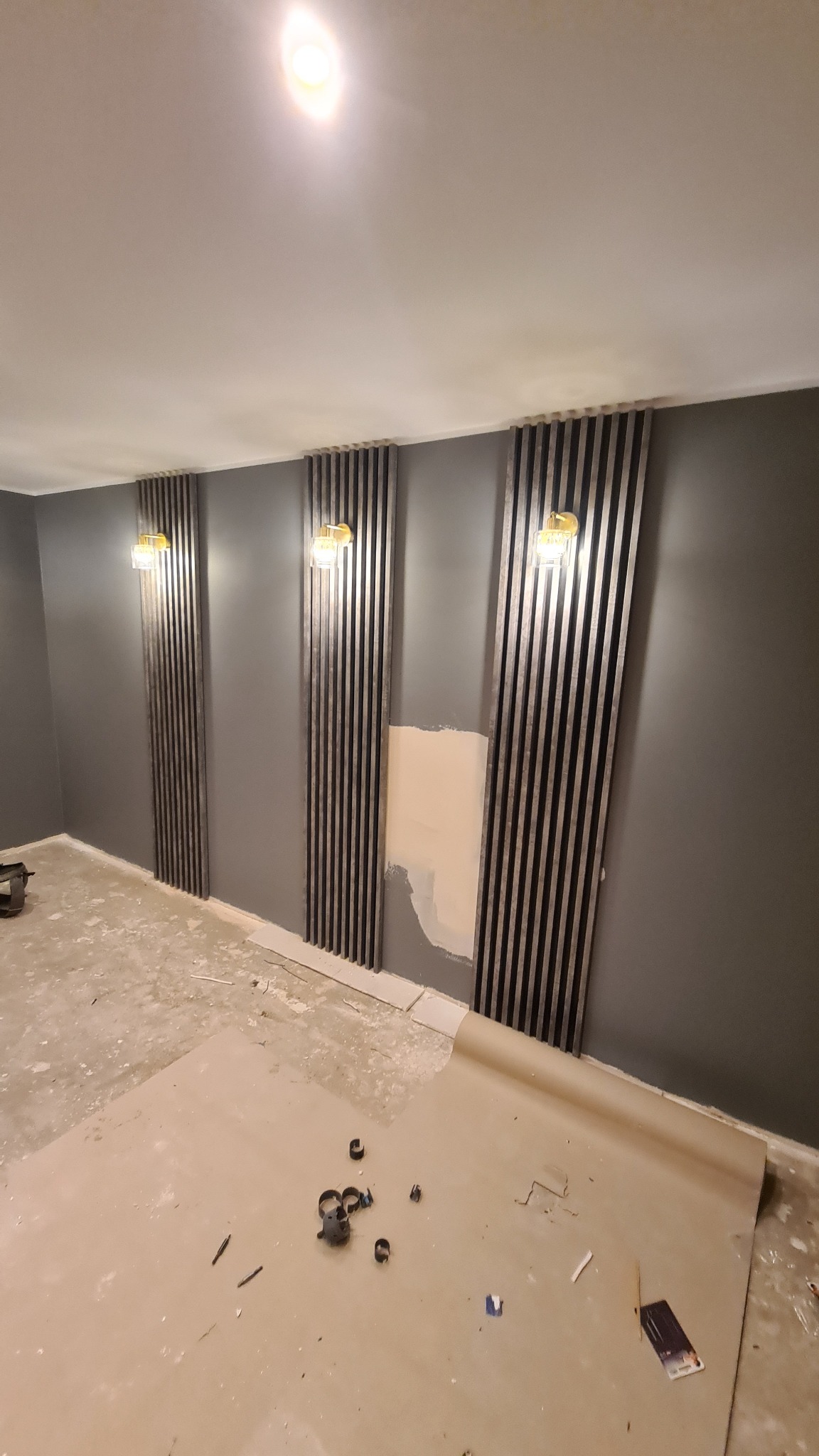 Szare ściany z dekoracyjnymi, pionowymi panelami z oświetleniem, fragment ściany z widocznym niedokończonym fragmentem, podłoga zabezpieczona papierem malarskim, widoczne narzędzia i akcesoria...