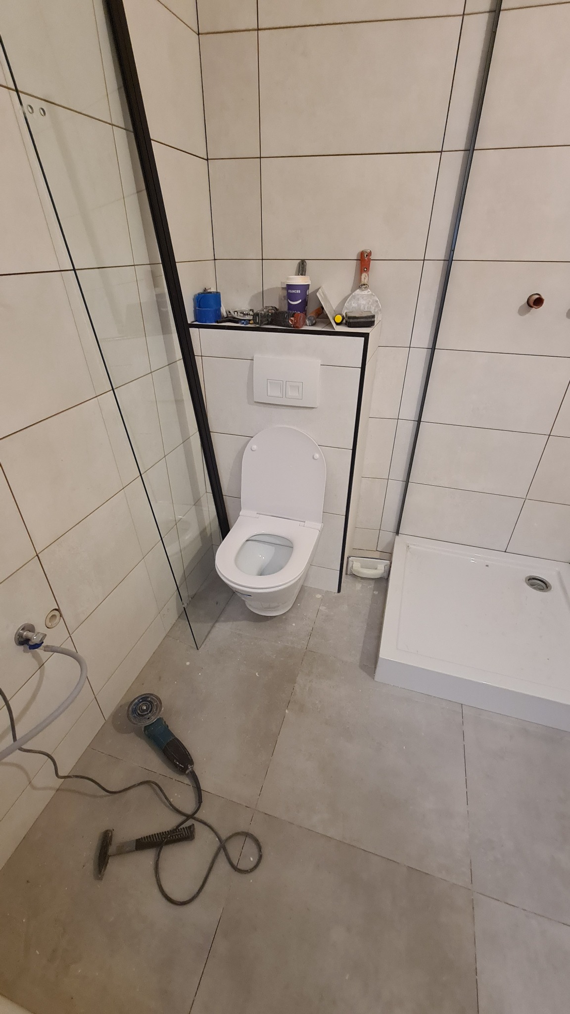 Biała toaleta podwieszana w trakcie montażu w łazience z ułożonymi płytkami, widoczne narzędzia: szlifierka kątowa i młotek, obok kabina prysznicowa z brodzikiem