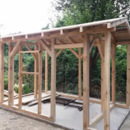 Drewniana konstrukcja zadaszenia w trakcie budowy, widoczne słupy, belki i elementy dachu, otoczenie z zielenią.