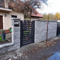 Nowoczesne ogrodzenie z szarej kostki łupanej z czarną, metalową furtką, prowadzące do domu z beżową elewacją i brązowymi drzwiami, widoczne jesienne liście na ziemi.