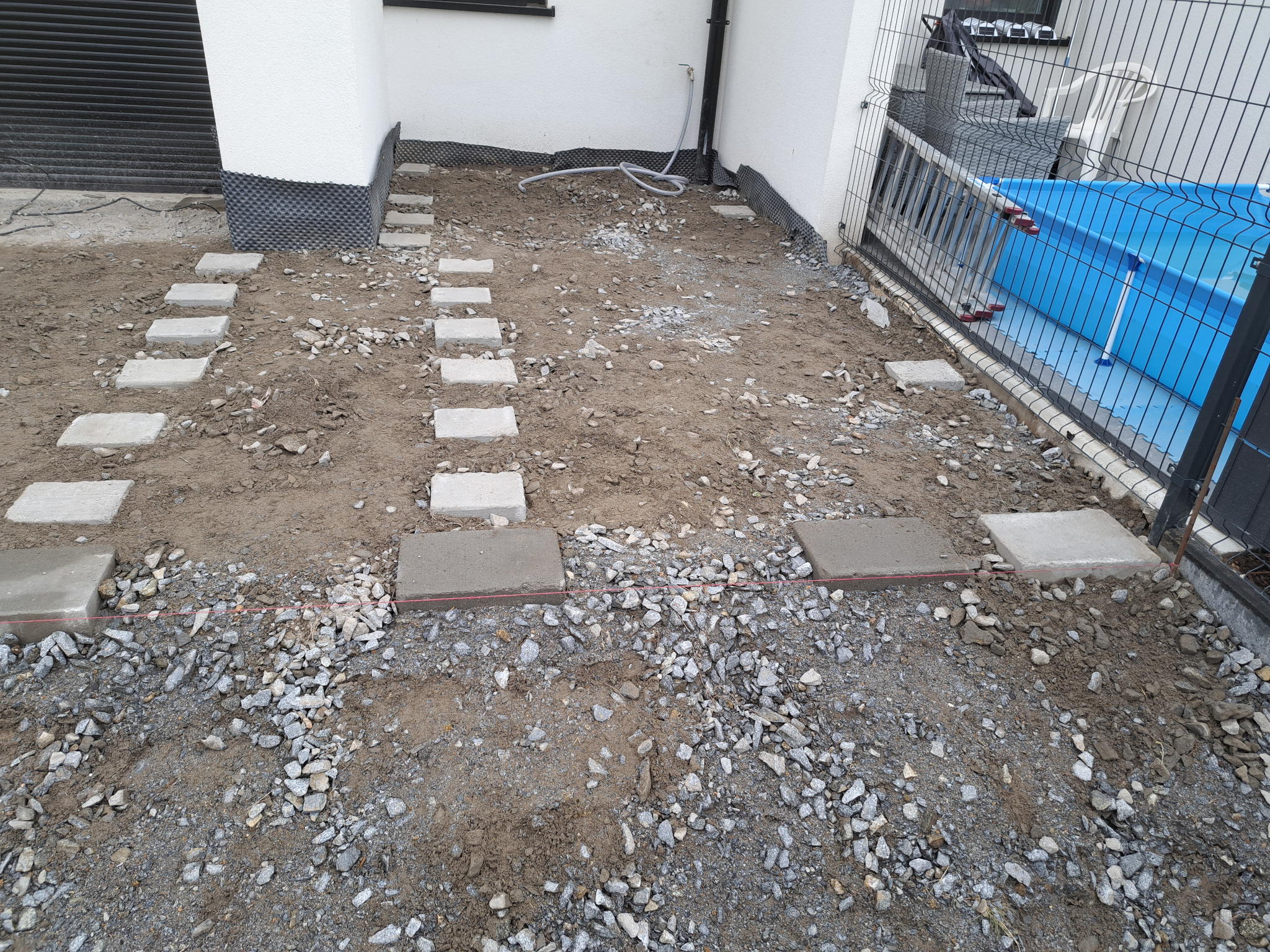 Przygotowanie podłoża pod taras lub ścieżkę z ułożonymi betonowymi płytami na podsypce żwirowej, widoczne napięte sznurki wyznaczające poziom, w tle ściana budynku i częściowo widoczny basen ogrodowy.