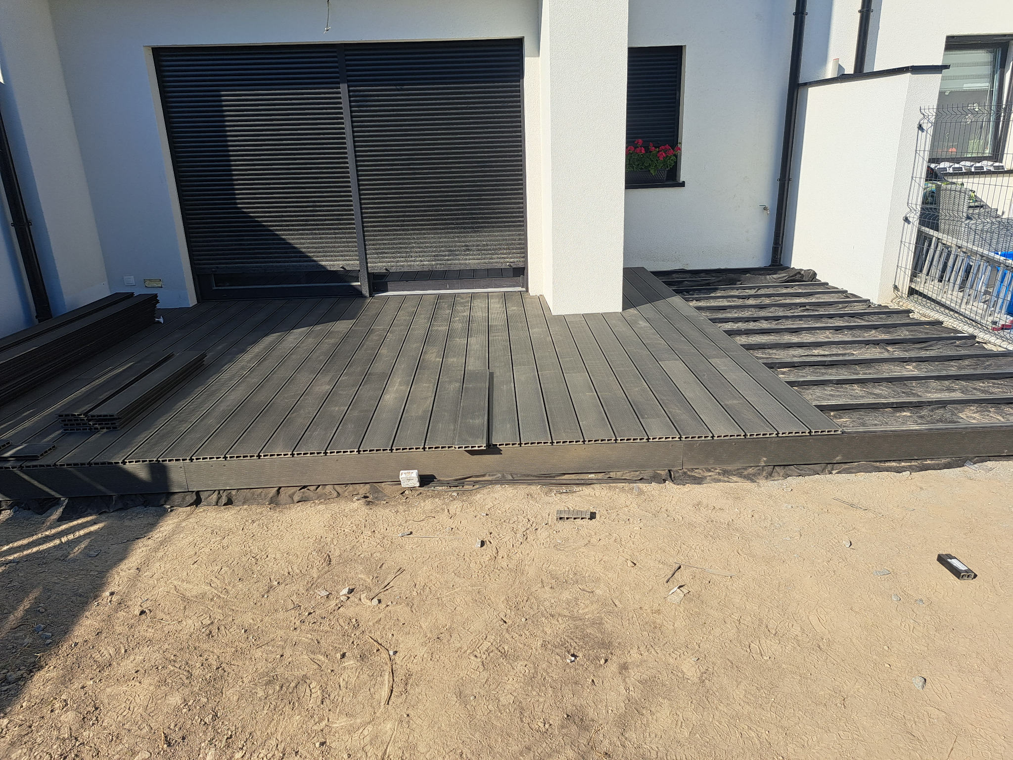 Montaż kompozytowej deski tarasowej na balkonie domu jednorodzinnego, widoczne legary, częściowo ułożona podłoga i stos desek. Dom z roletami zewnętrznymi i kwiatami w oknie.