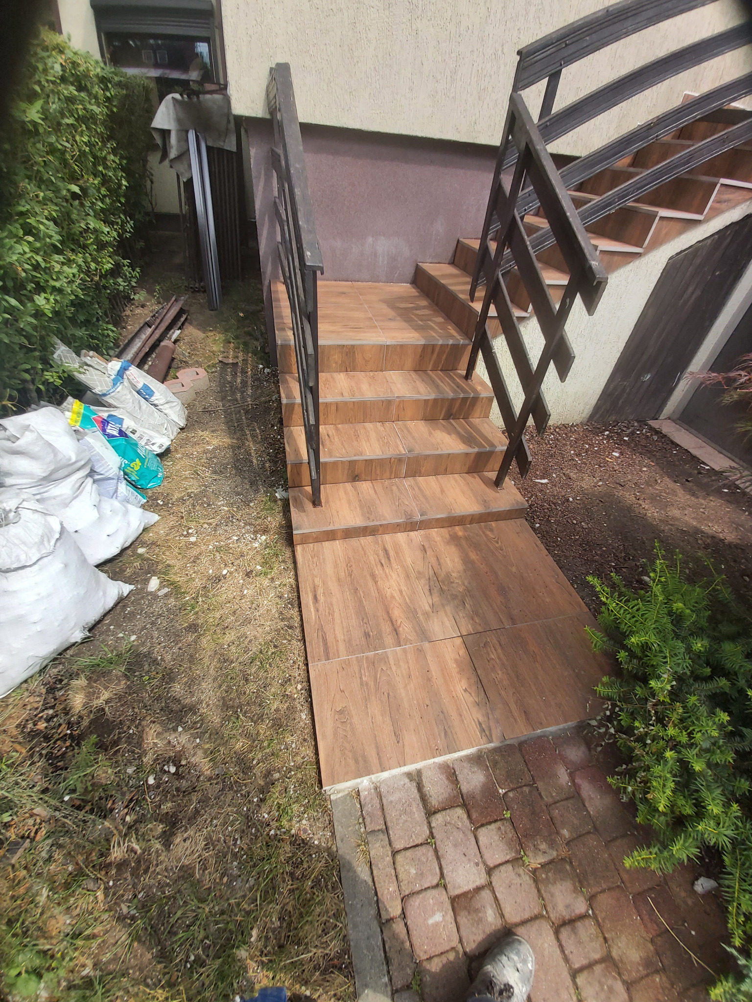 Wyłożone płytkami schody zewnętrzne z metalową barierką, prowadzące do domu, obok widoczne worki z materiałami budowlanymi i gruz.