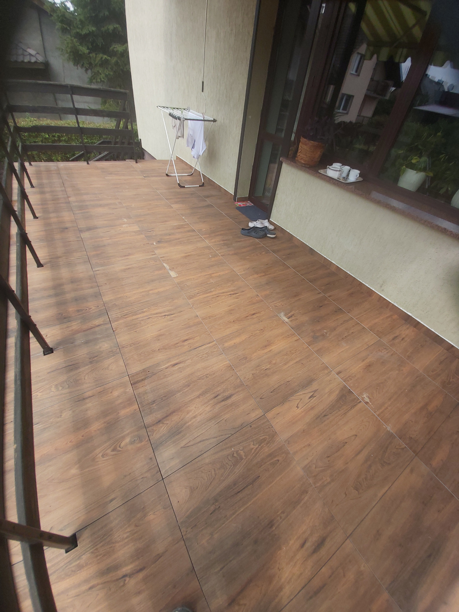 Wyłożony brązowymi płytkami balkon z metalową balustradą, suszarką na pranie i parą butów przy oknie.