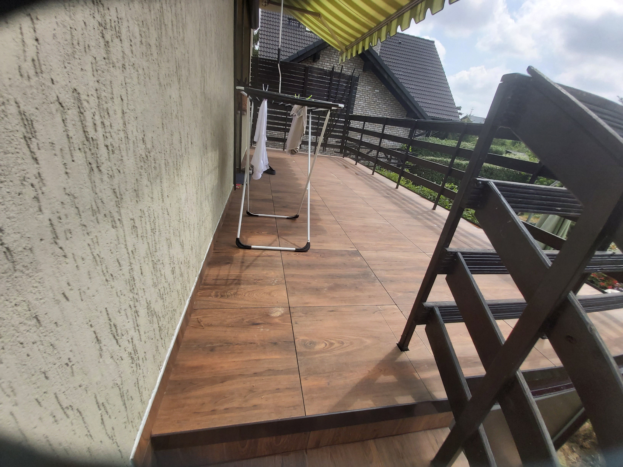 Balkon z drewnopodobnymi płytkami, szarym tynkiem strukturalnym na ścianie i rozkładaną suszarką na pranie, widok z góry na zewnątrz.