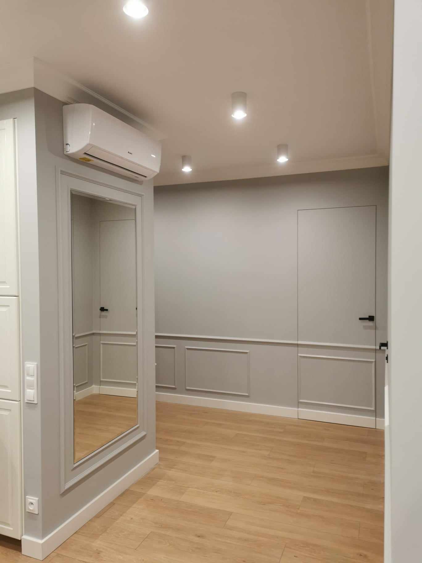 Jasne wnętrze z panelami podłogowymi, szarą ścianą z dekoracyjnymi panelami, ukrytymi drzwiami z czarną klamką, lustrem w ozdobnej ramie i klimatyzatorem.