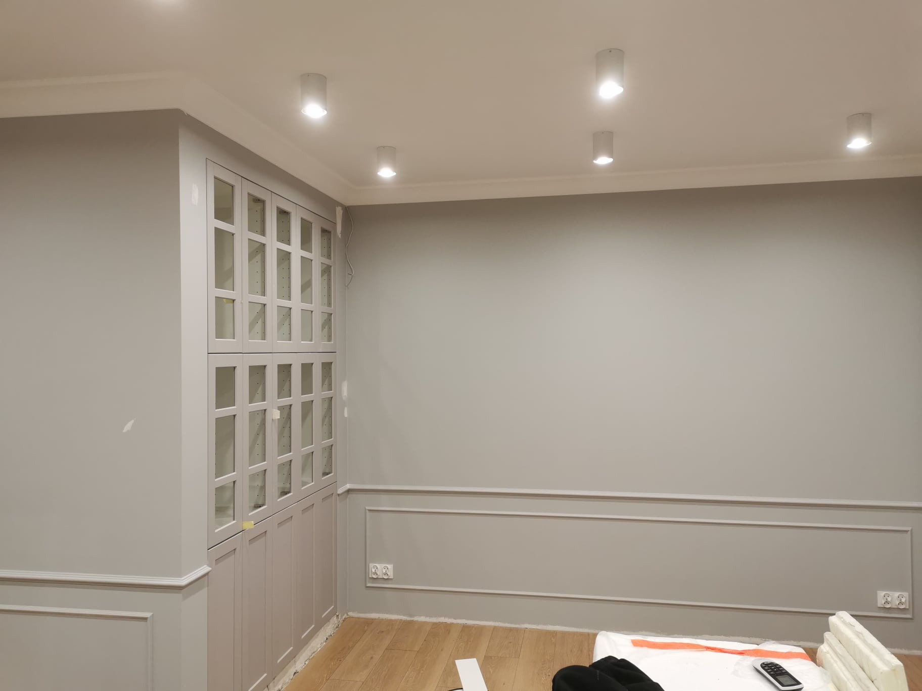 Szare wnętrze po remoncie, z zabudowaną szafą z przeszklonymi drzwiczkami i listwami dekoracyjnymi na ścianach, oświetlone punktowymi lampami sufitowymi, z widocznymi elementami zabezpieczeń podłogi.