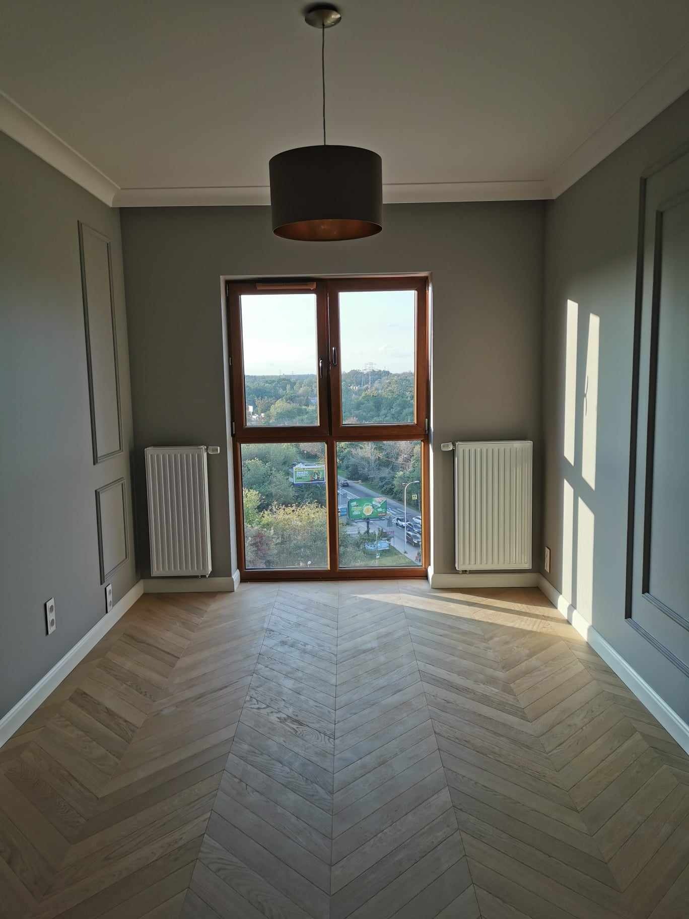Pusty pokój z parkietem ułożonym w jodełkę, szare ściany z dekoracyjnymi panelami, okno z widokiem na miasto i wisząca lampa z ciemnym abażurem.