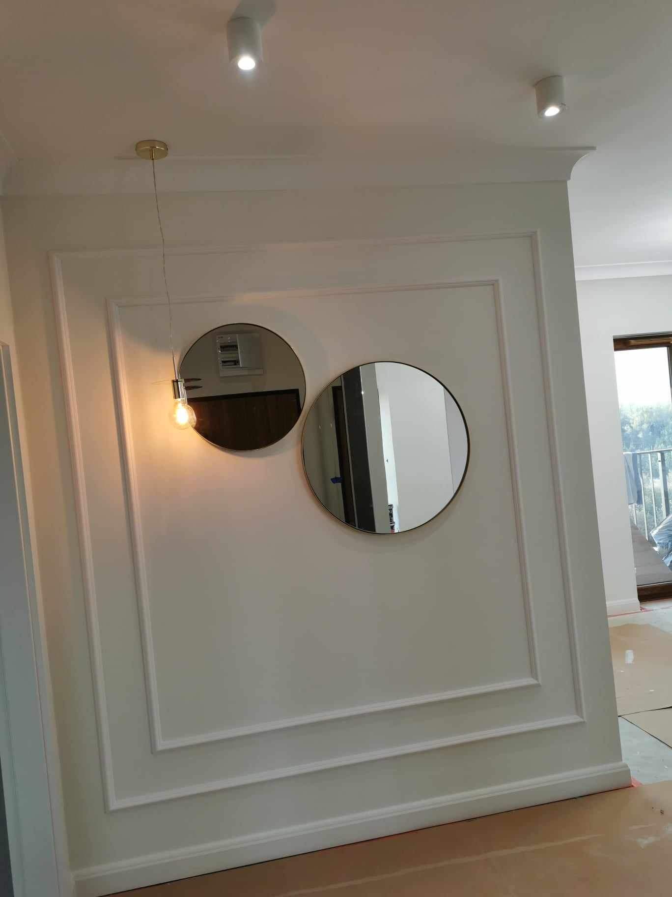 Biała ściana z dekoracyjnymi listwami i dwoma okrągłymi lustrami, jedno z widoczną żarówką na kablu, w tle widoczne drzwi balkonowe.