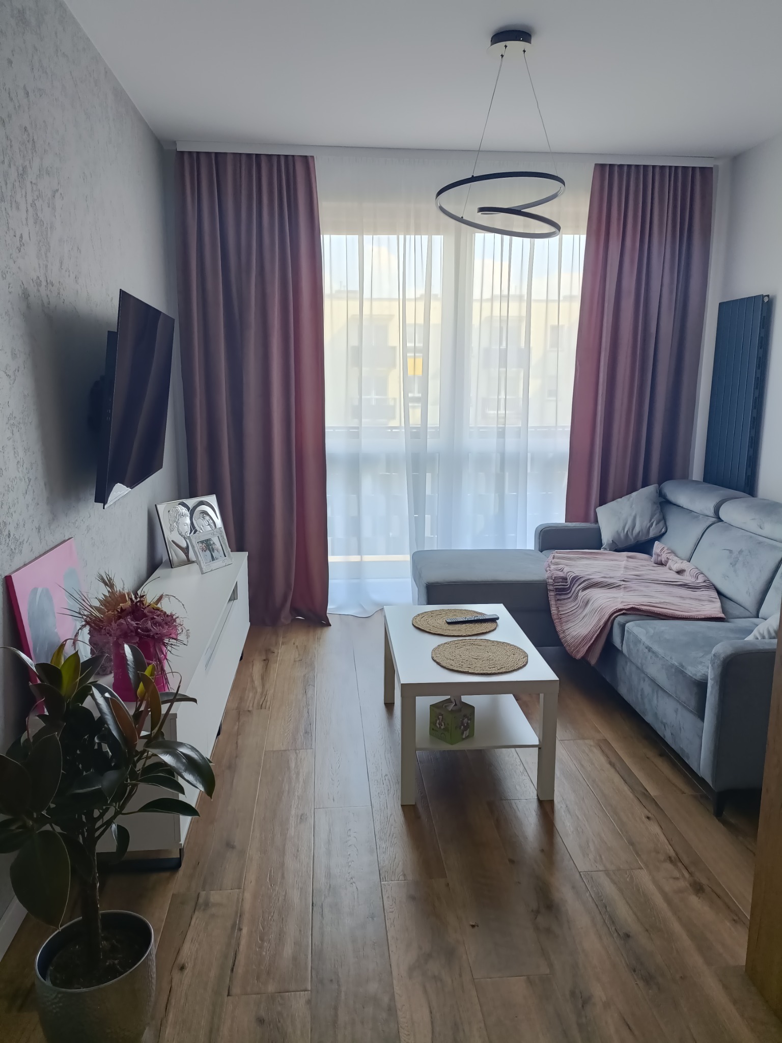 Salon z drewnianą podłogą, szarą sofą z pledem, białym stolikiem kawowym, telewizorem na ścianie z dekoracyjną fakturą i nowoczesną lampą sufitową