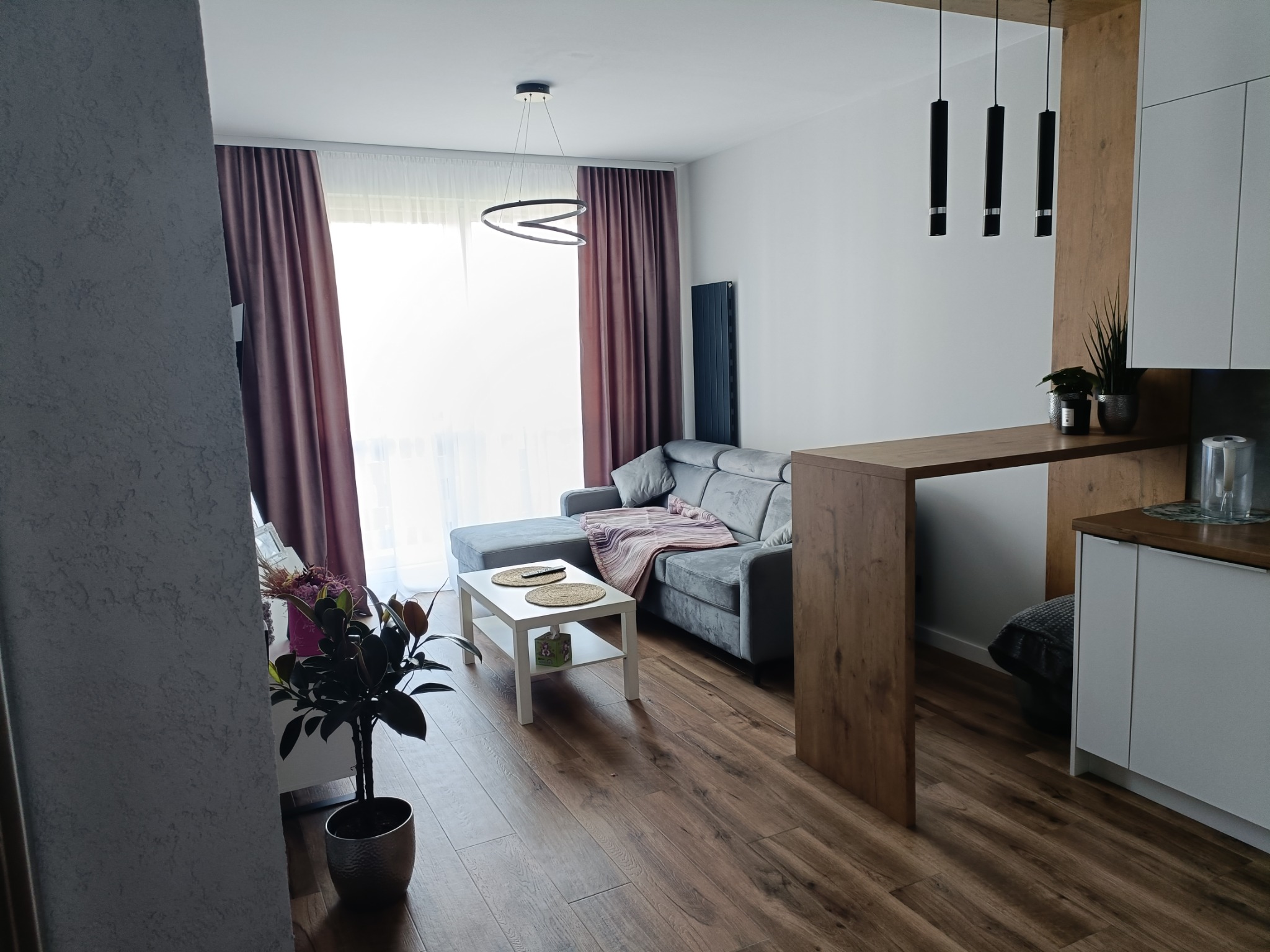 Nowoczesne wnętrze salonu z szarym narożnikiem, drewnianą podłogą, białym stolikiem kawowym i dekoracjami roślinnymi, oświetlone wiszącymi lampami.