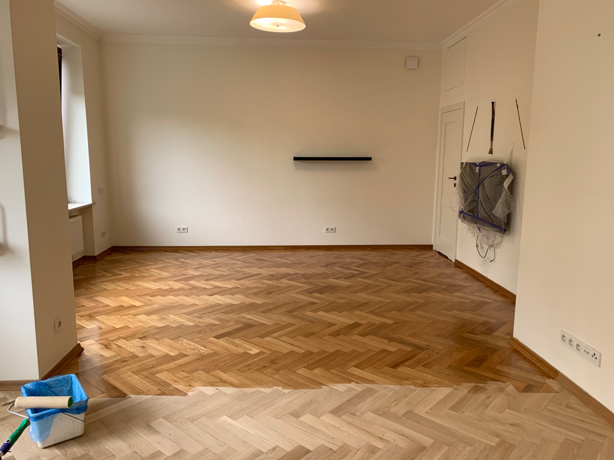 Pokój z parkietem w jodełkę, częściowo odnowionym, widoczny wałek malarski w wiadrze, ściany przygotowane do malowania.