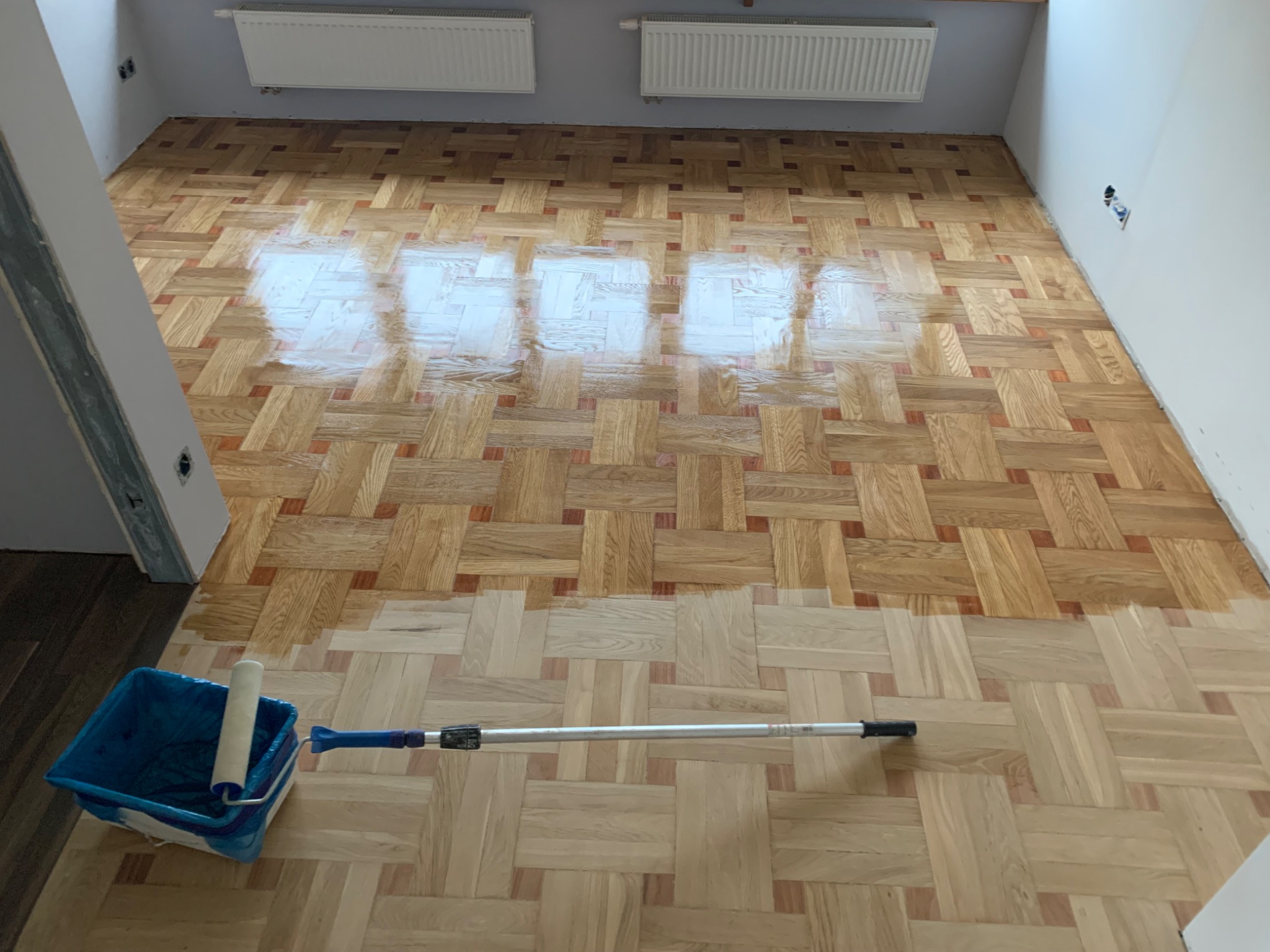Parkiet ułożony w jodełkę, częściowo pokryty lakierem, z widocznym wałkiem malarskim i kuwetą na podłodze, w tle grzejniki na ścianie.