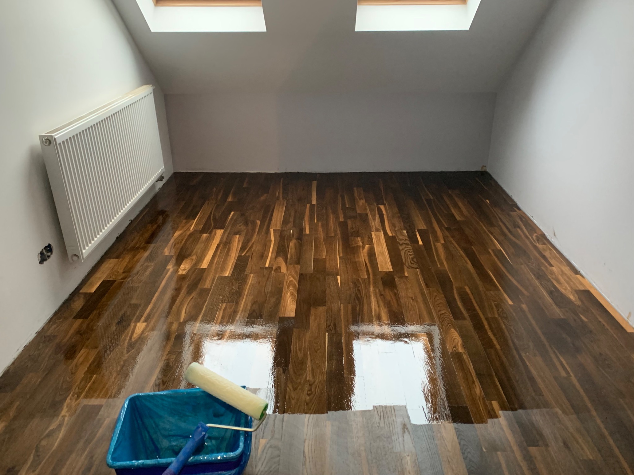 Świeżo polakierowana, ciemna drewniana podłoga w pokoju na poddaszu, z widocznym wałkiem do lakierowania i niebieskim wiadrem, odbijająca światło z okien dachowych.
