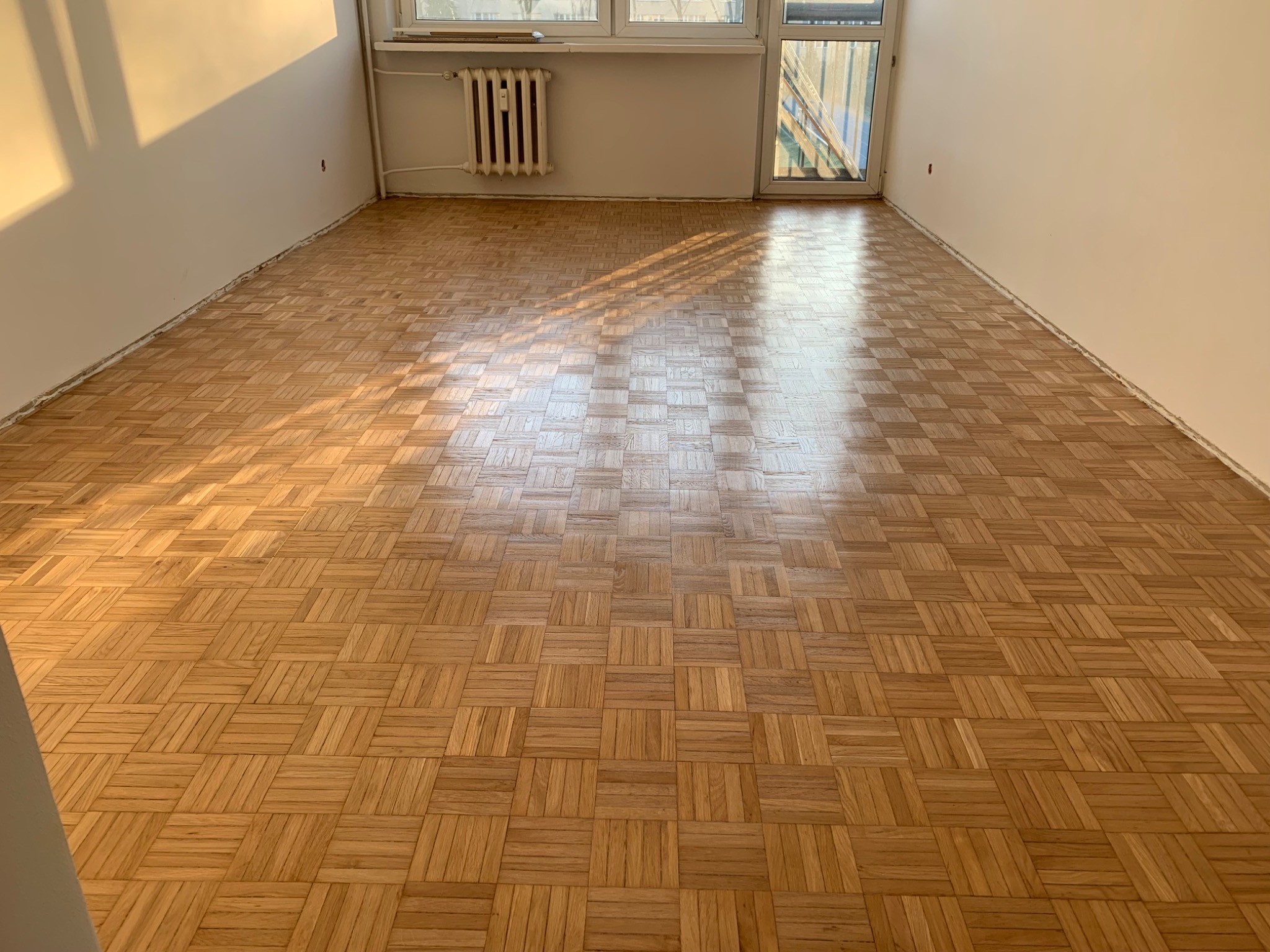 Parkiet ułożony w kwadraty, świeżo po renowacji, widoczne ślady po listwach przypodłogowych, wpadające promienie słoneczne przez okno balkonowe, grzejnik żeliwny pod oknem.