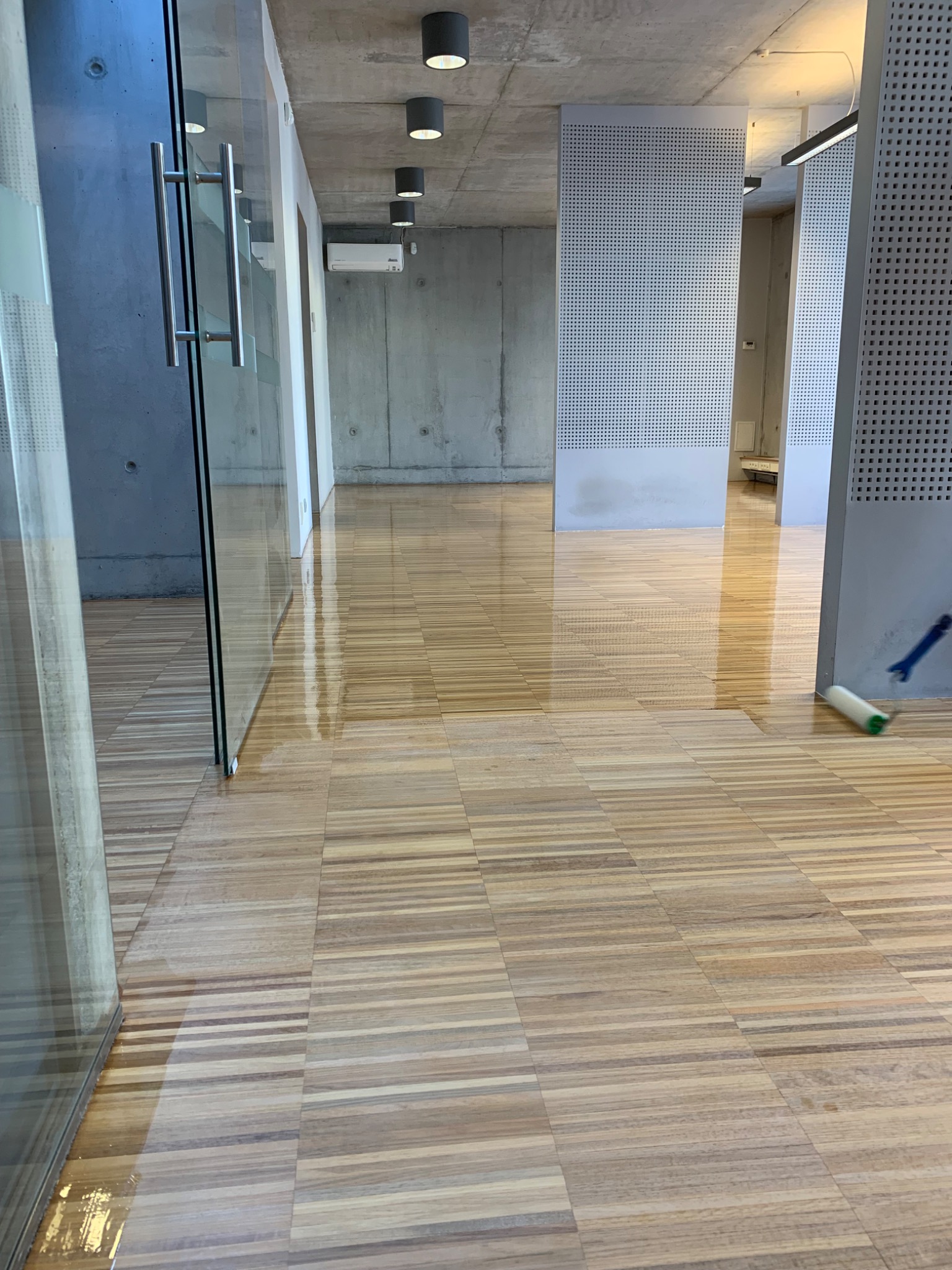 Cyklinowanie parkietu w nowoczesnym biurze z betonowymi ścianami i szklanymi drzwiami, widoczny wałek malarski w trakcie pracy.