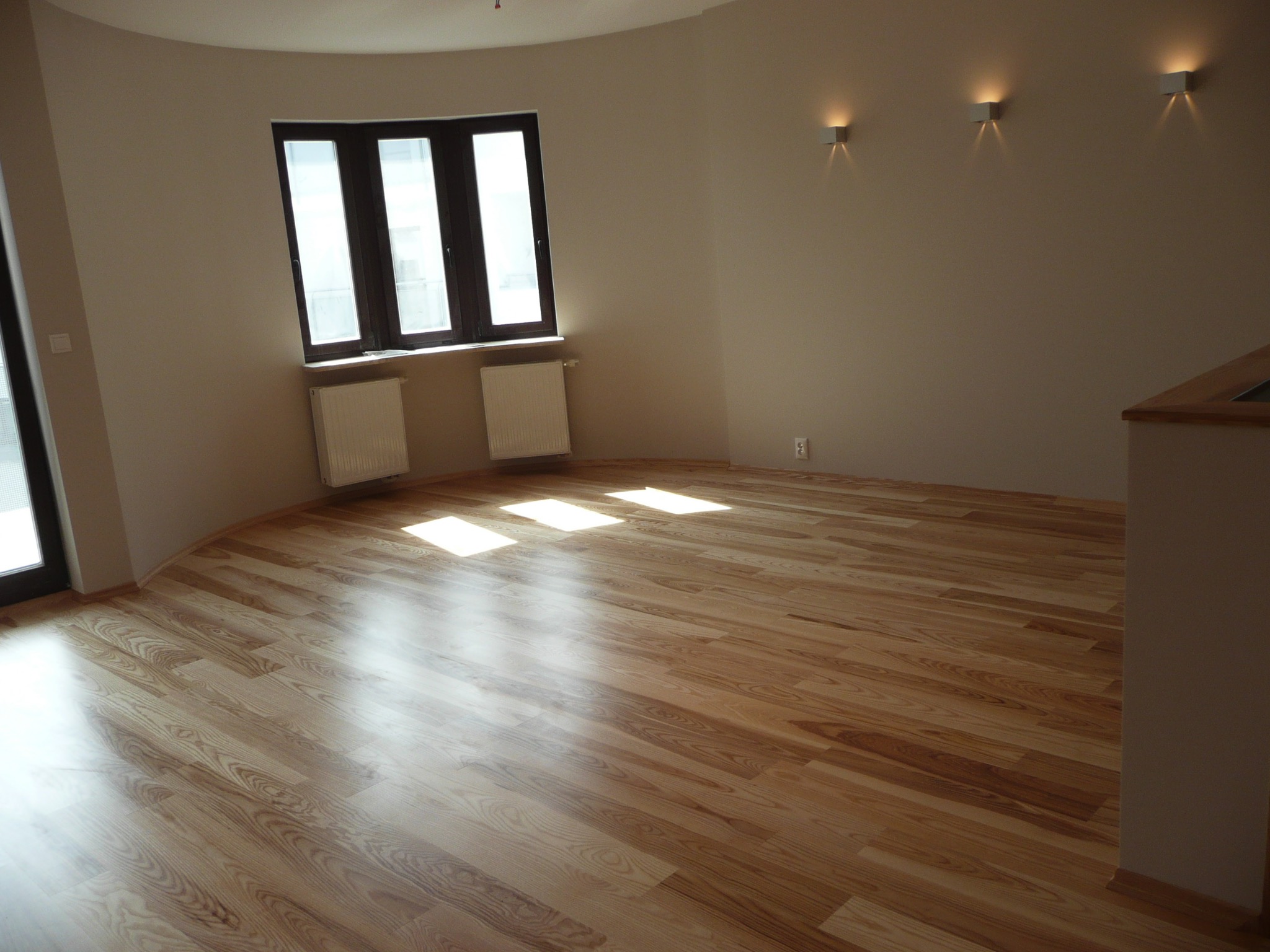 Jasne, drewniane panele podłogowe ułożone w przestronnym, jasnym pokoju z zaokrągloną ścianą, oświetlonymi kinkietami i oknem z grzejnikami, przez które wpadają promienie słoneczne.